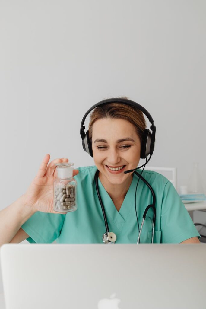 Nurse in green scrubs wearing headphones smiles while holding a pill bottle during a video call.