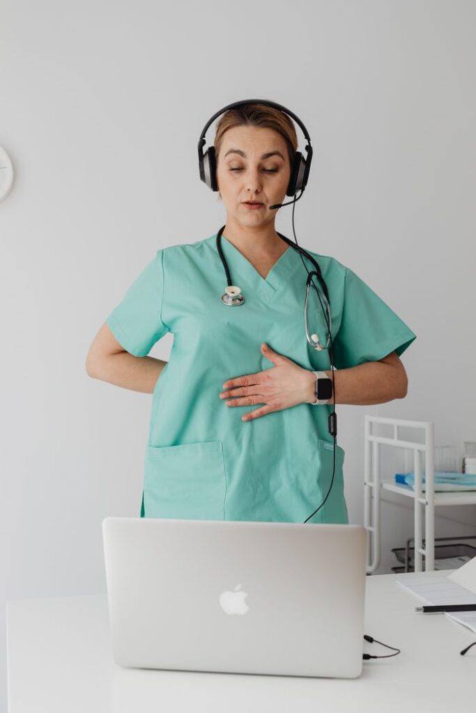 Medical professional in scrubs using a laptop for online consultation with a headset.